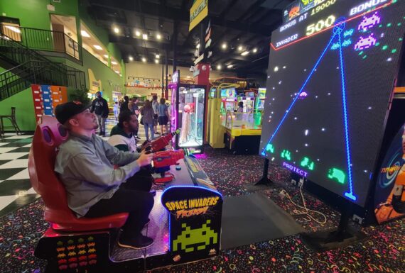 Two individuals playing a large arcade game of Space Invaders in a vibrant gaming center, surrounded by other arcade machines and colorful decor.